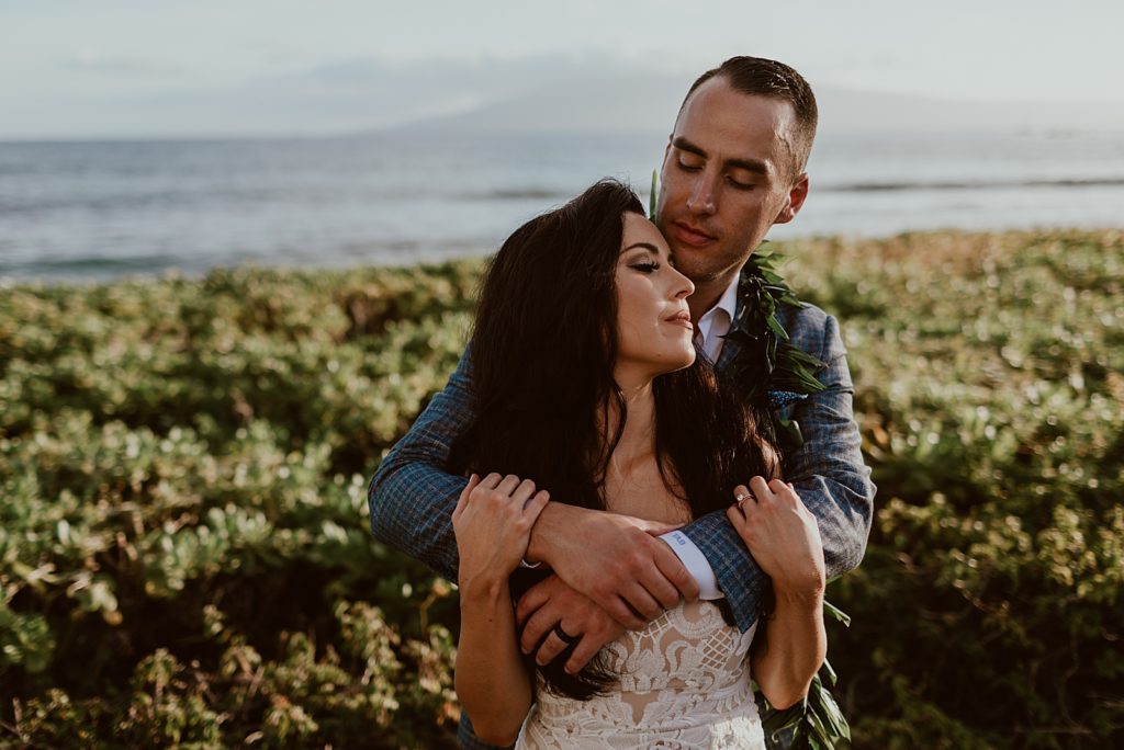 Groom holding Bride from behind in front of the ocean