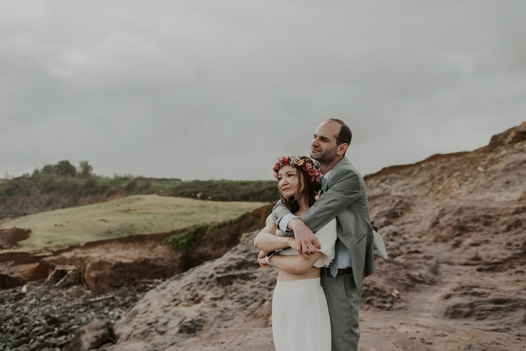 Groom hugging Bride from behind looking out at the ocean