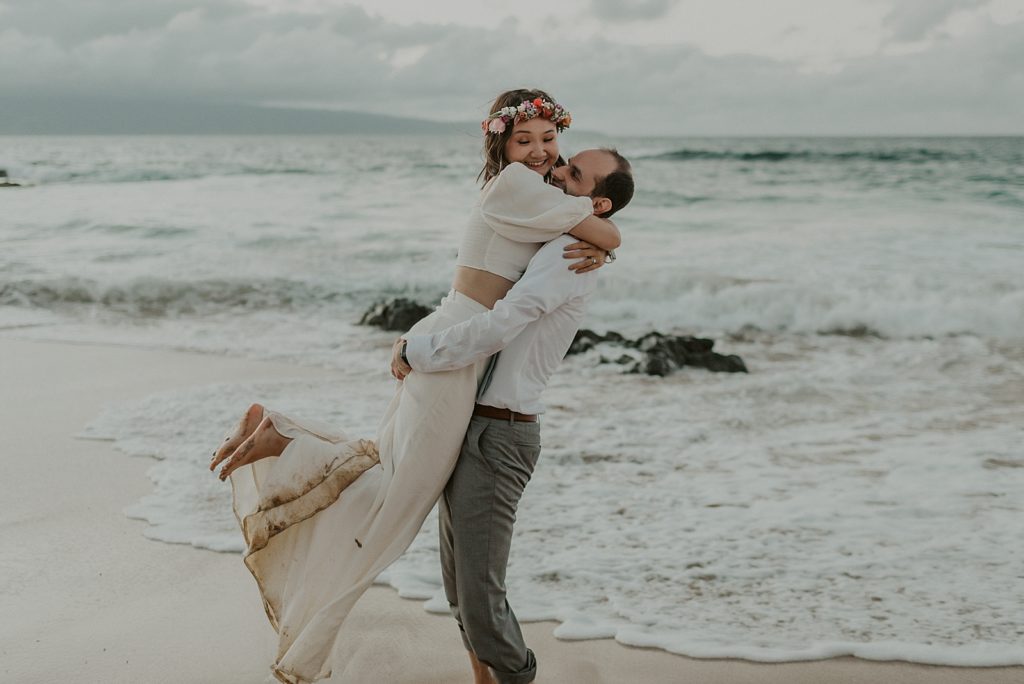 Groom lifting Bride up on sand in ocean wave coming in