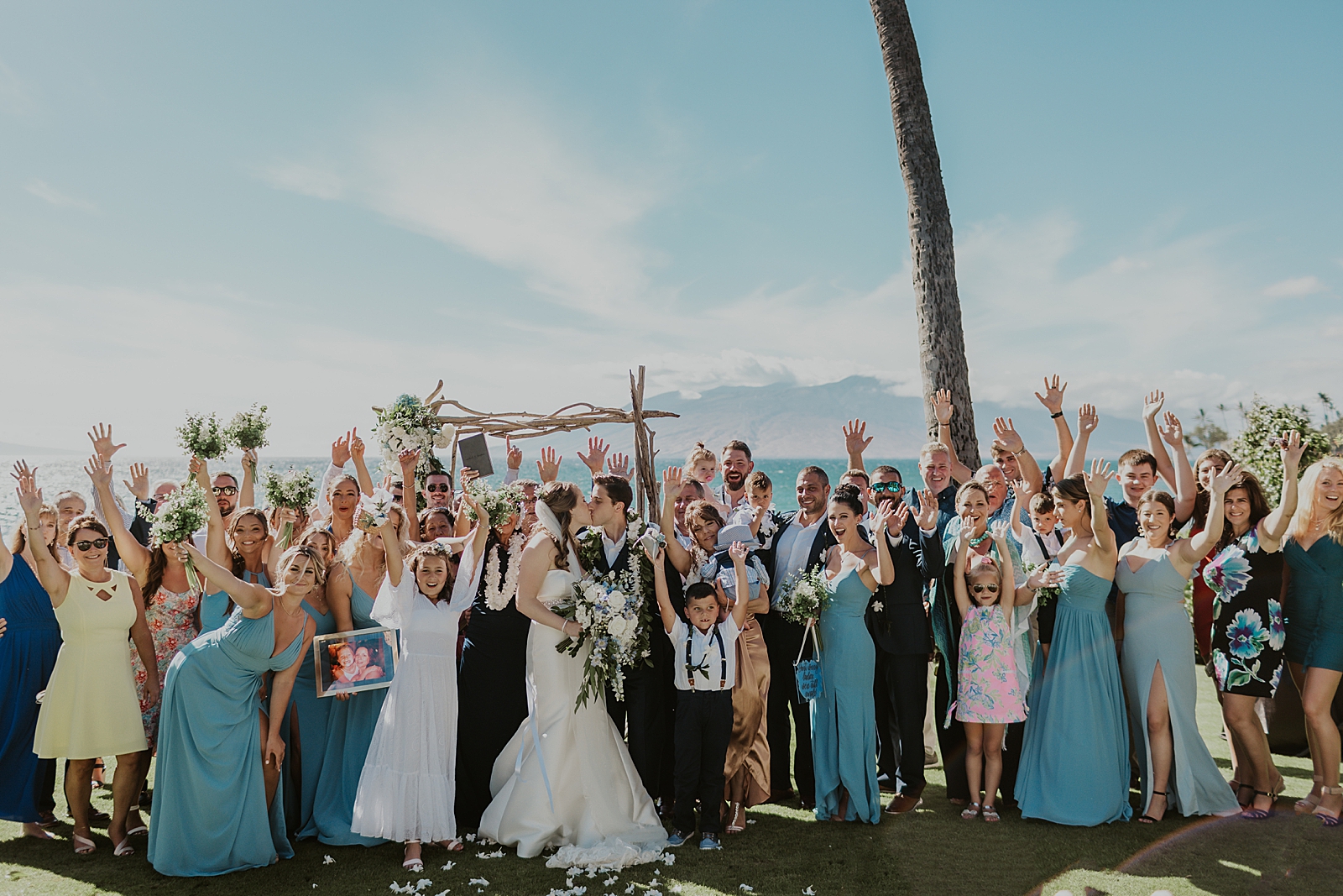 Portrait of wedding guests holding hands up as Bride and Groom kiss outside by the water