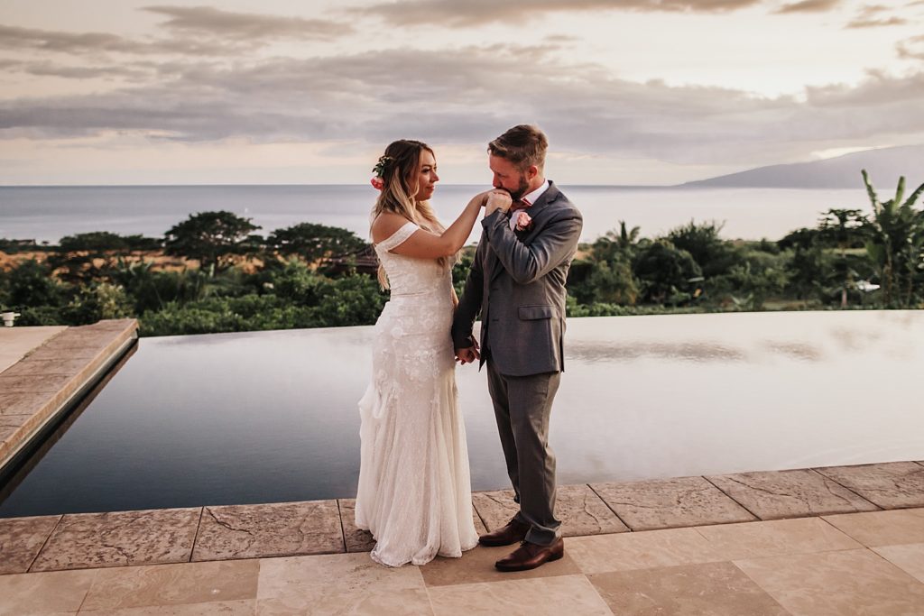 Groom kissing Bride on the hand by the pool as the sun sets