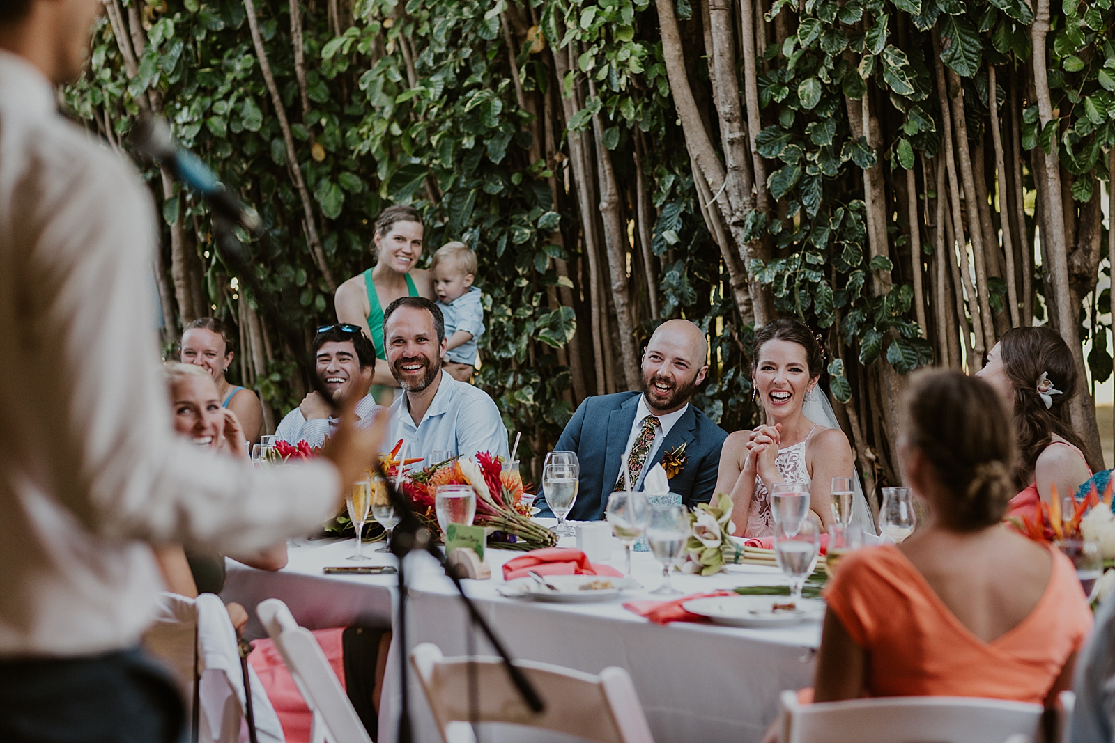 Shot of Bride and Groom sitting with guests at long Reception table listening to speech
