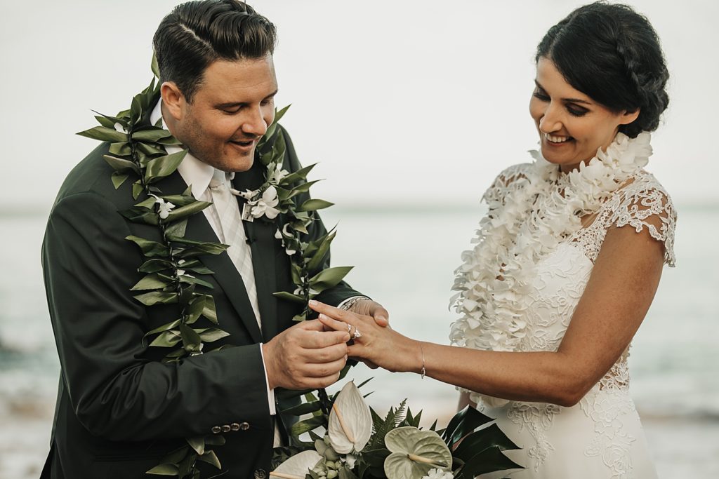 Groom putting wedding band on Bride's finger by the ocean