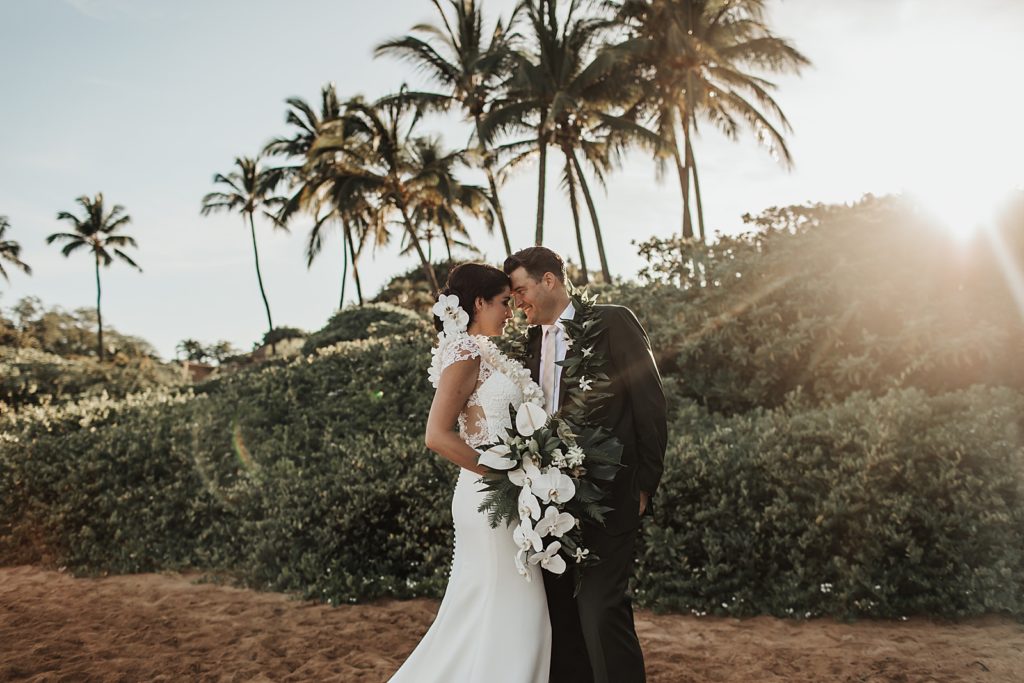 Bride and Groom resting their heads on each other standing on the sand with the sun setting